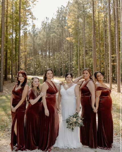 Bride in white wedding gown standing in the middle of five bridesmaids wearing burgundy dresses outdoors with trees behind them. Bride is holding a bouqet.