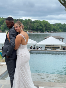The bride is wearing a white wedding gown holding the groom in a gray suit from behind him. There is white tents pictured in the background, along with a body of water.