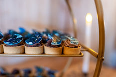 Small cakes with blue details are pictured. There is multiple sitting on a gold table with white candles lit in the background.
