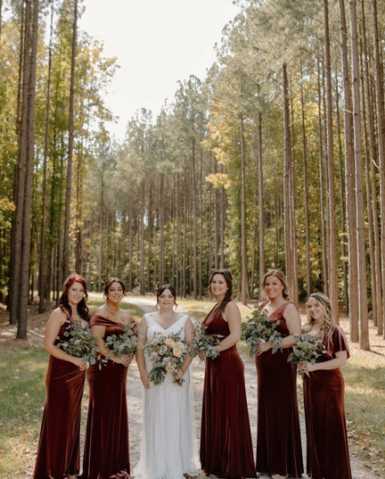 Bride in the center of 5 bridesmaids wearing burgundy gowns on a dirt road with trees in the background. Everyone is holding bouqets.