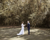 A bride in a white gown reads from a small book in front of a groom in a black suit. There is a forest behind them. 