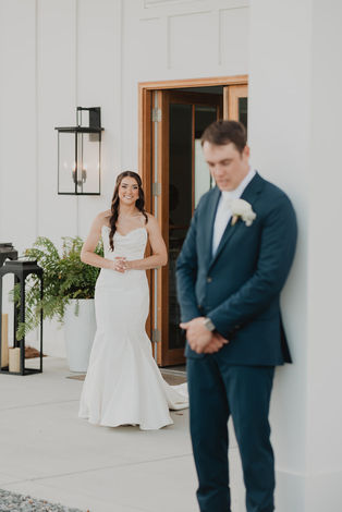 A bride stands behind the groom. He stands in front of a white post and she looks from behind him.
