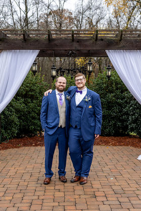 Two men in blue suits pose together. There is a wooden bridge behind them with white curtains hanging off.