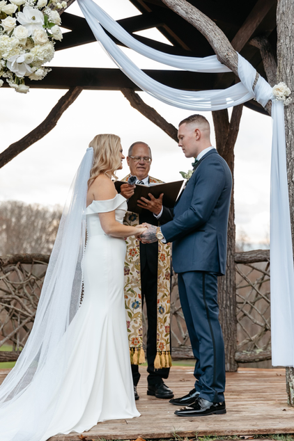 Wedding couple in wedding attire standing at the alter. The ceremony is outside under a wooden arbor.