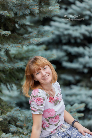 Smiling woman with floral print blouse and skirt posing next to trees. Slava Photo