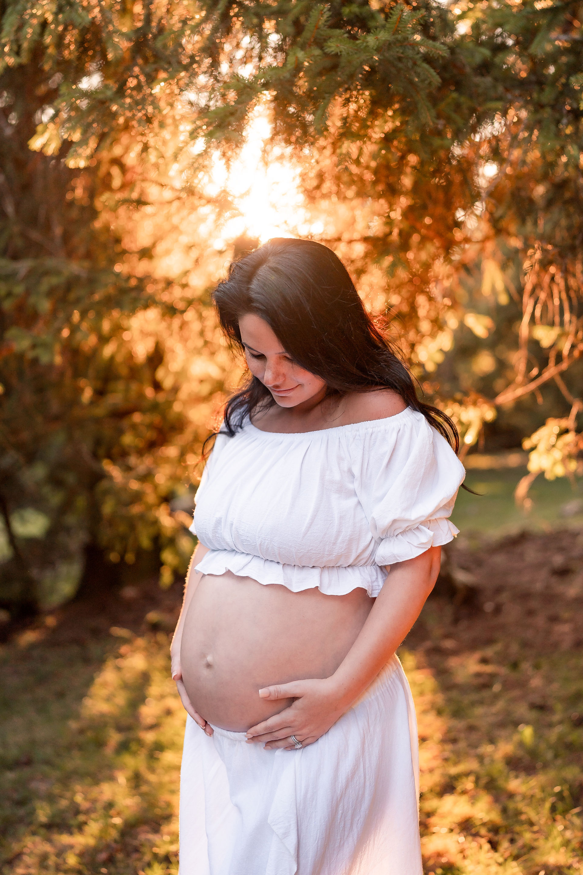 Pregnant woman in white dress holds belly, standing in sunlit forest. Warm sunset glow filters through trees, creating serene mood.