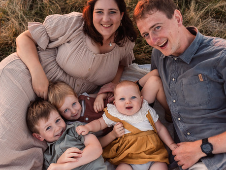 Family of five smiling, lying on grass. Baby in yellow, others in neutral tones. Warm, relaxed outdoor scene.