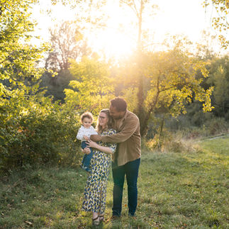 Family enjoying a sunny day outdoors. A smiling man embraces a woman holding a child. Greenery and sunlight in the background.