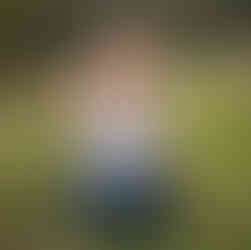 Smiling toddler in white shirt and jeans is sitting on the ground, playing with a flower. Sun is setting behind him giving the scene an idyllic feel.