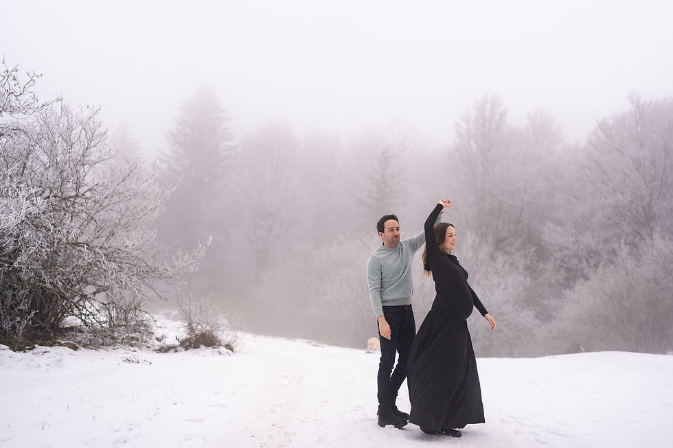 Pregnant woman dances with a man in a snowy forest. Foggy background, trees covered in frost. She wears a black dress; he wears a gray sweater.
