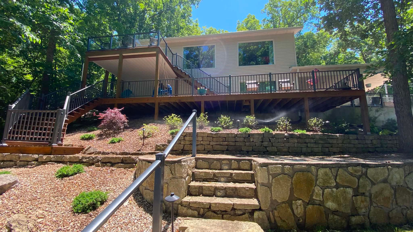 Lake home backyard landscaping with tiered retaining walls, stone steps, and elevated deck overlooking wooded surroundings.
