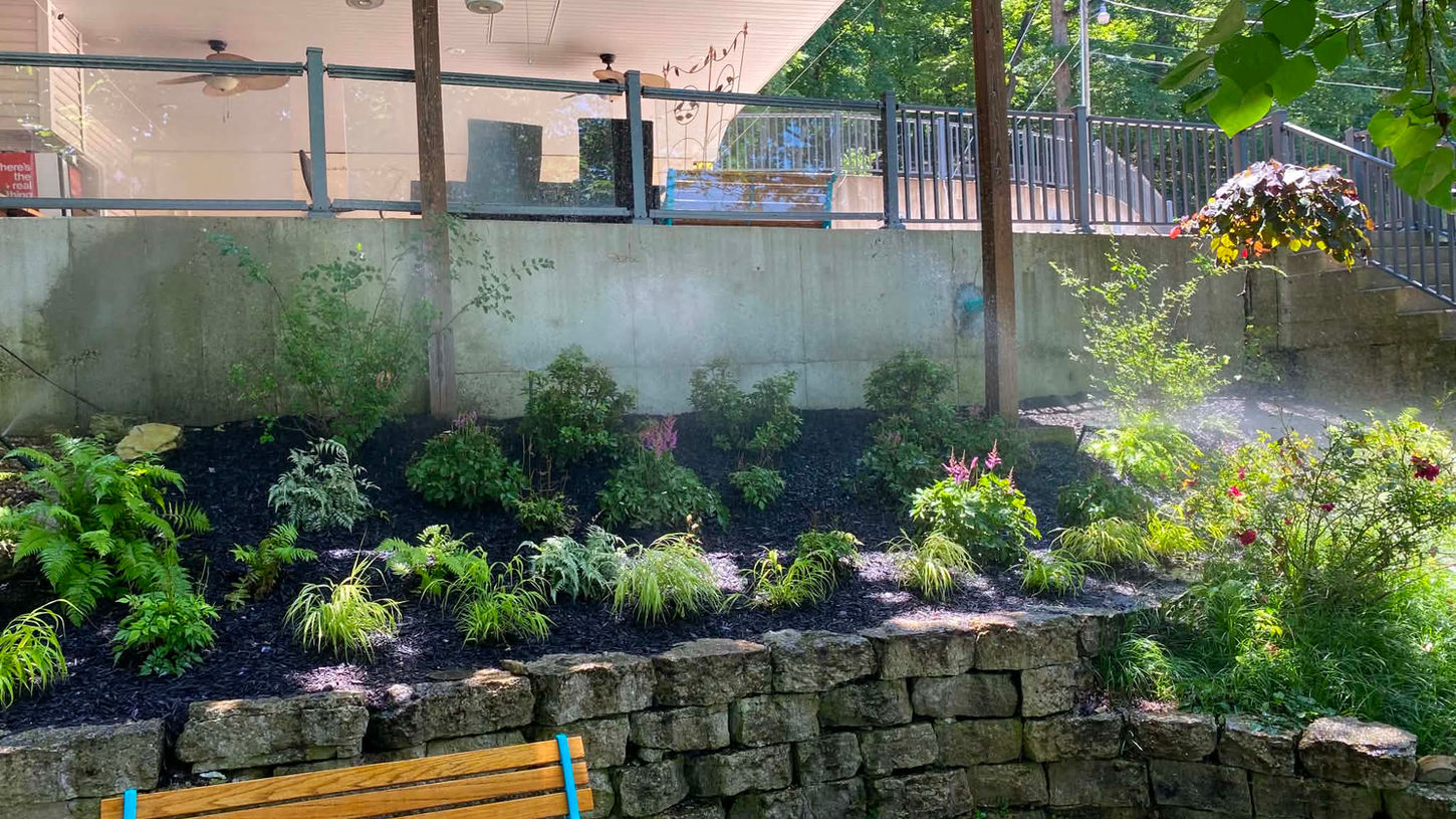 Low-maintenance shade landscaping with mulch beds, ornamental grasses, and retaining wall design under a raised deck.
