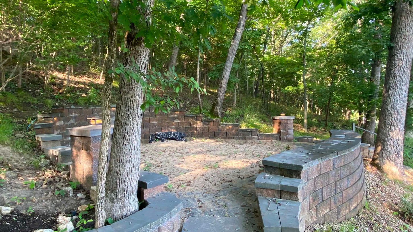 Custom hardscape design with curved retaining walls, stone steps, and natural wooded landscaping for outdoor living space.