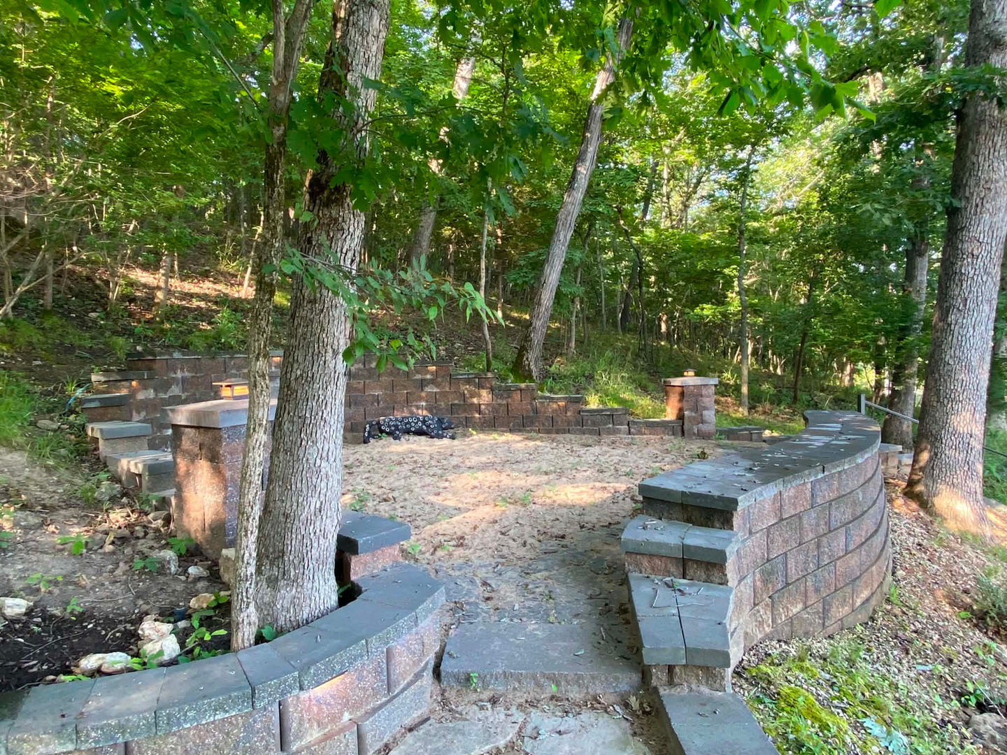 Custom hardscape design with curved retaining walls, stone steps, and natural wooded landscaping for outdoor living space.