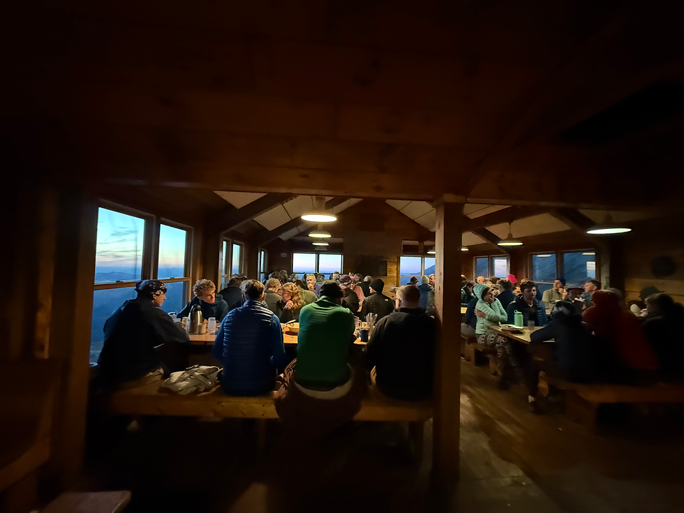 Hikers eating a family-style dinner at long tables with benches at Lakes of the Clouds Hut. The sun is setting with. mountain views through the windows.