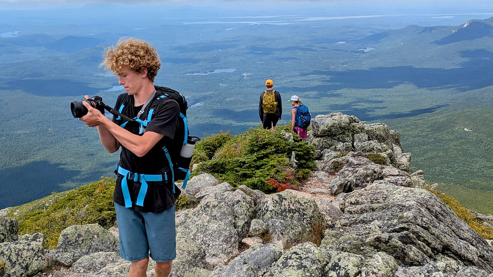 Mom and her teenage sons, descending Katahdin's Hunt Trail. They are standing on a rocky cliff and one boy is holding a camera. The blue sky has puffy clouds forming shadows on the green valley below.