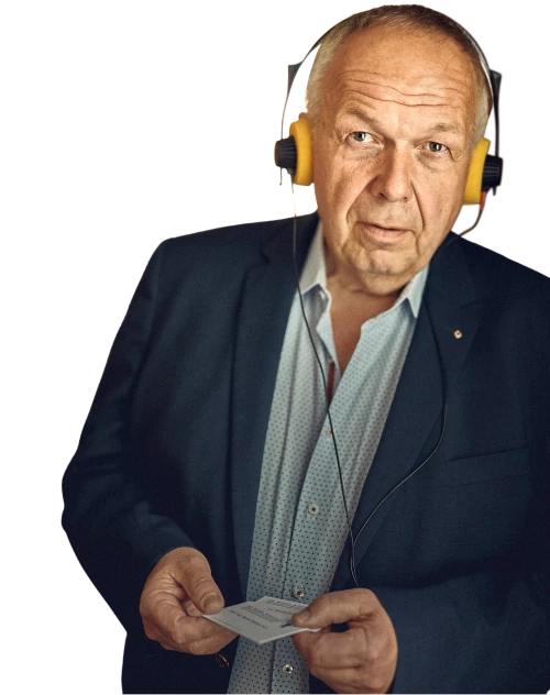 Ein älterer Mann mit Glatze und einem kleinen Lächeln trägt einen dunklen Anzug mit einem gepunkteten Hemd ohne Krawatte. Er hält eine Karte in der Hand und hat gelbe Kopfhörer über den Ohren.