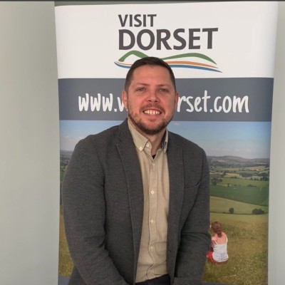 James Maidment stands in front of a Visit Dorset banner, smiling, wearing a beige collared shirt and brown jacket