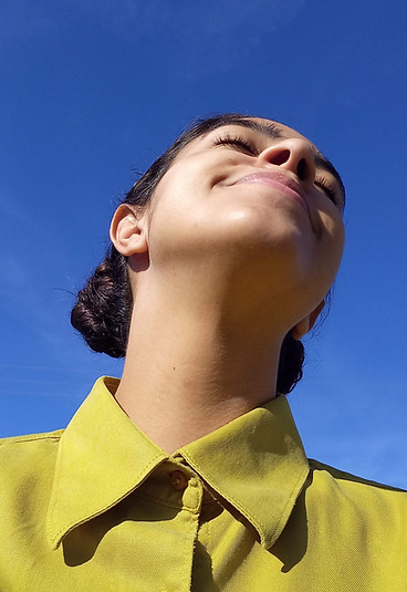 Mariana com o cabelo preso olhando para o céu. Ela está vestida com uma blusa verde e o céu está azul
