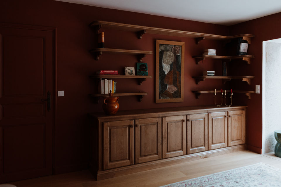 Cette bibliothèque a été conçue comme un élément architectural à part entière, structurant le mur et définissant l’identité de l’espace. Le dessin sur mesure associe des rangements bas fermés à des étagères ouvertes, permettant un équilibre entre fonctionnalité et mise en scène décorative.
La teinte profonde des murs, volontairement enveloppante, met en valeur le bois et renforce la sensation de profondeur. Les proportions, le rythme régulier des étagères et les consoles apparentes s’inspirent du vocabulaire classique, réinterprété de manière sobre et contemporaine.
L’intégration d’œuvres, de livres et d’objets sélectionnés avec retenue transforme la bibliothèque en support narratif, participant à l’atmosphère calme du lieu.