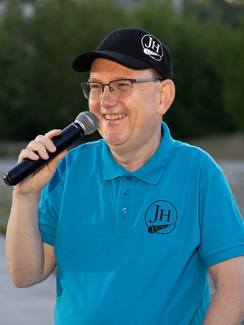 Ein lächelnder Moderator spricht mit Mikrofon bei einem Event im Freien.