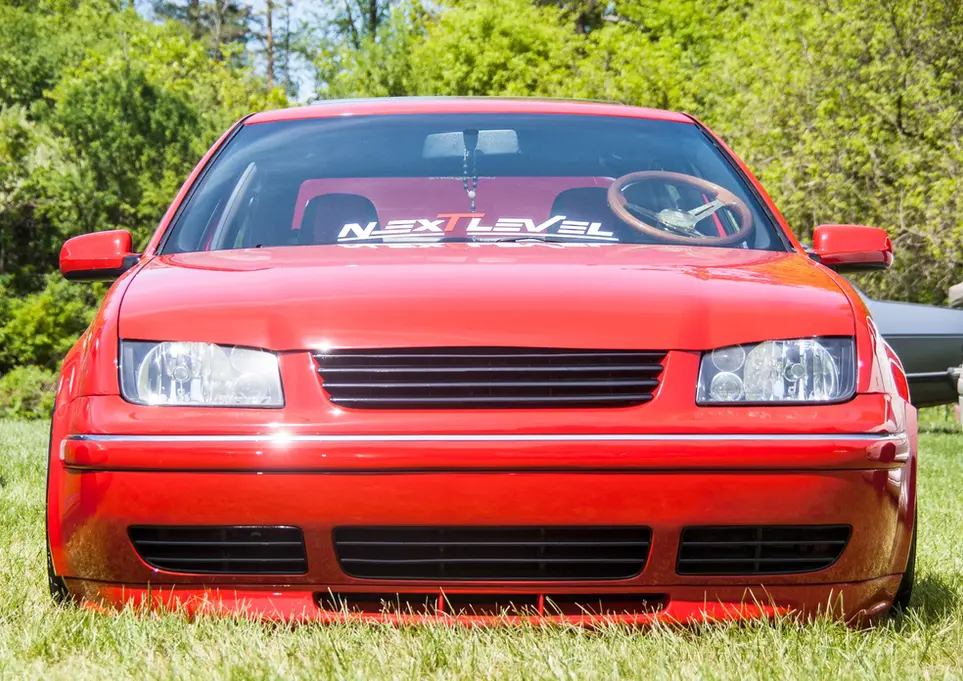 Red lowered Volkswagen Jetta with "NEXT LEVEL" banner on grass.