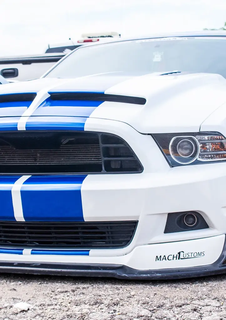 White Ford Mustang with blue racing stripes, "MACH" spoiler, parked outdoors.