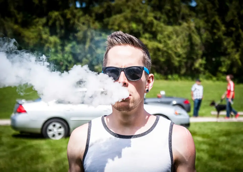 Man in sunglasses exhaling large vape cloud, outdoor gathering.