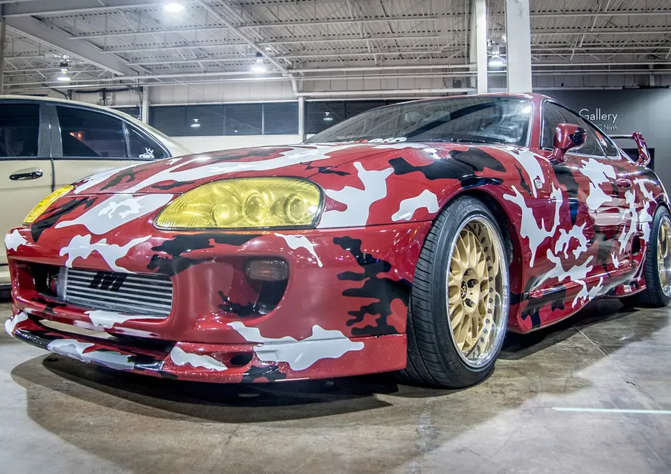 Red and white camouflage Toyota Supra with gold wheels at car show.