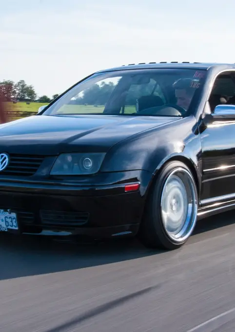 Black Volkswagen Jetta driving on road with license plate visible and background.