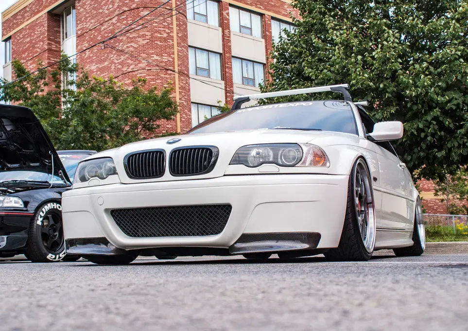 White BMW E46 sedan parked outdoors, brick building background.