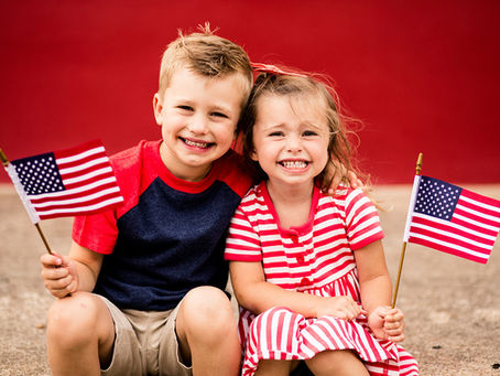 two smiling children holding American flags while sitting on a sidewalk