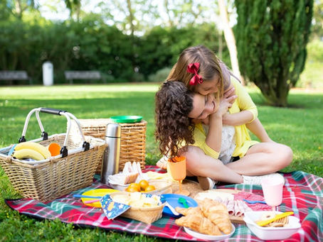 a young daughter hugging her mother on a picnic blanket outside surrounded by grass and food