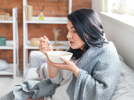 a young woman eating from a bowl of soup while sitting on a sofa while wrapped in a blanket