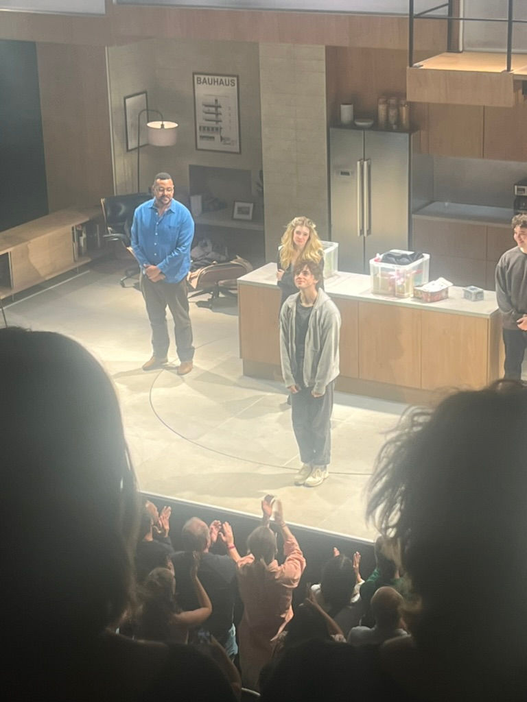 Three people stand on stage, smiling, in a modern kitchen set as the audience applauds. A "BAUHAUS" poster is visible on the wall.