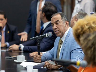 Altair Moraes de terno azul claro, falando ao microfone durante em reunião na Assembléia, sentado à mesa com outros participantes ao fundo.