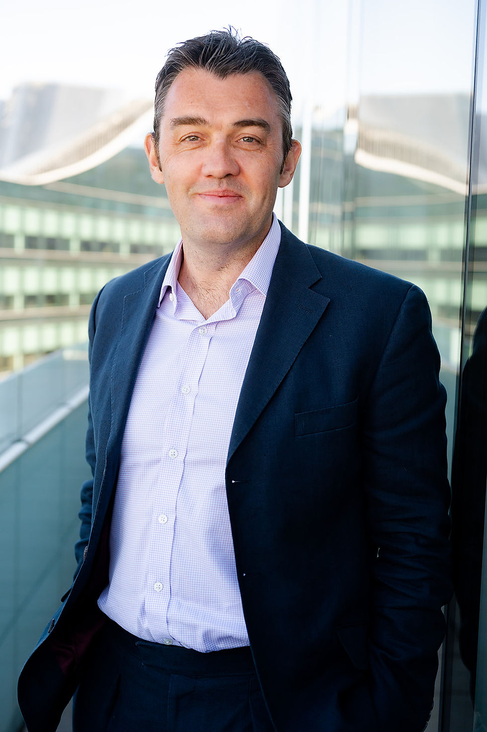 Smiling man in a navy blazer and light check shirt stands by a glass office building.