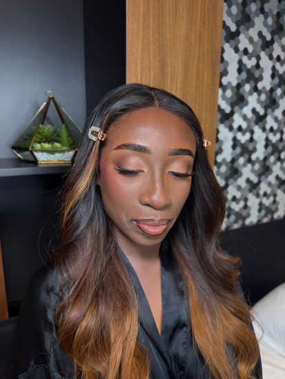 Soft glam makeup on a dark-skinned client, featuring warm neutral eyeshadow, defined lashes, and a glossy nude lip, with long waves pinned back during the glam process.