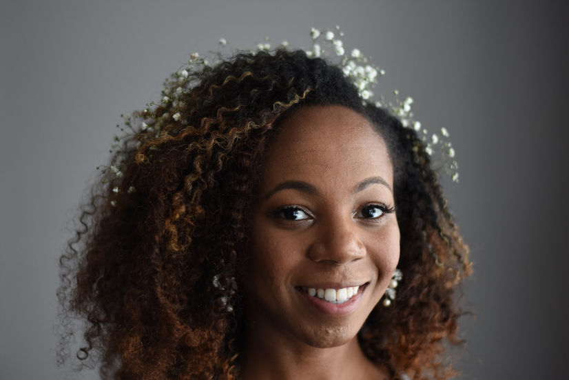 Smiling bride with curly natural hair and a delicate hairpiece, wearing soft glam bridal makeup against a neutral backdrop.