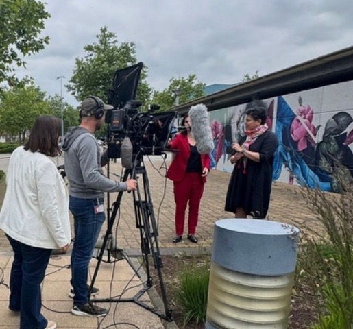 Outdoor film crew records two women near a vibrant building mural.