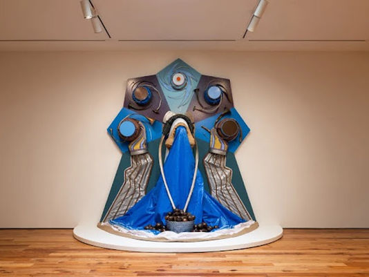 Abstract blue-and-gray sculpture on a gallery pedestal, with draped fabric and circular forms, in a minimalist room.