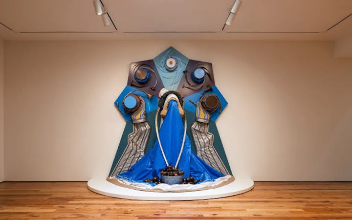 Abstract blue-and-gray sculpture on a gallery pedestal, with draped fabric and circular forms, in a minimalist room.