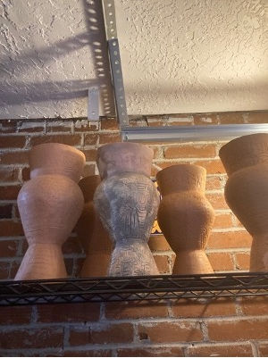 Four tall decorative vases on a metal shelf against a brick wall and ceiling, in muted pink, gray, and tan tones.