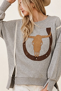 Close up of a model in a grey oversized sweatshirt showing raw-edge cream contrast seams, a longhorn graphic, and side-slit cuffs.
