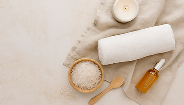 Flat lay of spa essentials including a wooden bowl of bath salts, rolled white towel, amber dropper bottle, and lit candle on a beige linen surface.