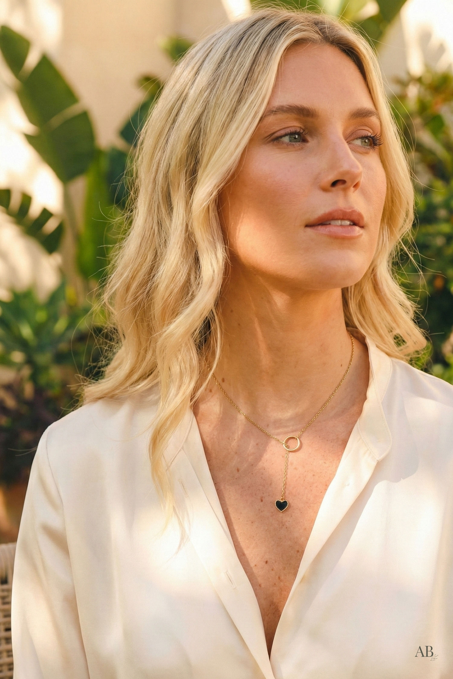 Sunlit lifestyle shot of a blonde model wearing a gold heart lariat necklace with a cream silk blouse.