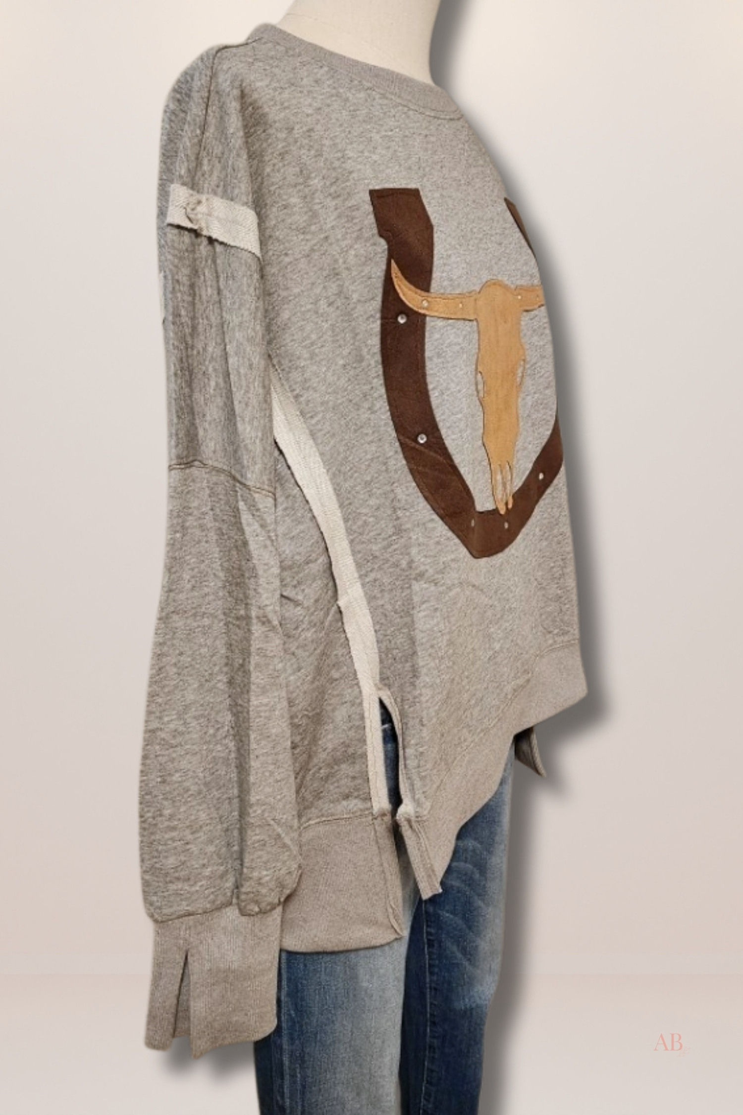 Side view of a grey graphic sweatshirt on a mannequin showing raw-edge cream seam details, side-slit cuffs, and hi-lo hem.