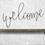 Thumbnail: Black wire 'Welcome' wall décor on rustic wood shelf against white textured wall, modern farmhouse style accent
