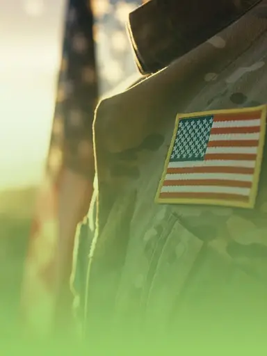 Soldier's back, American flag patch on military uniform, blurred flag background.