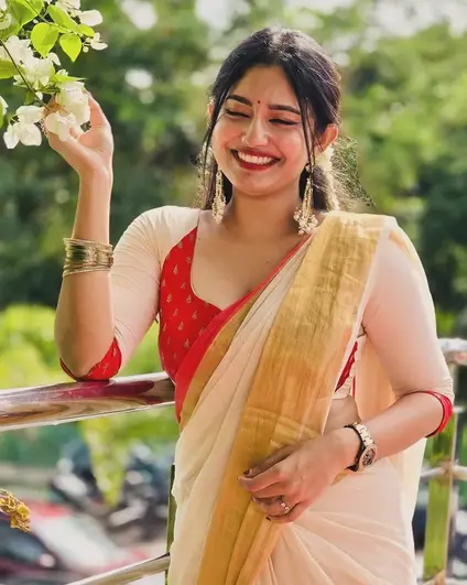 Nandita S. Cherian wearing a Kerala white saree with red border, round hanging jumkas, red lipstick, golden watch, and golden bangles, showcasing elegance.